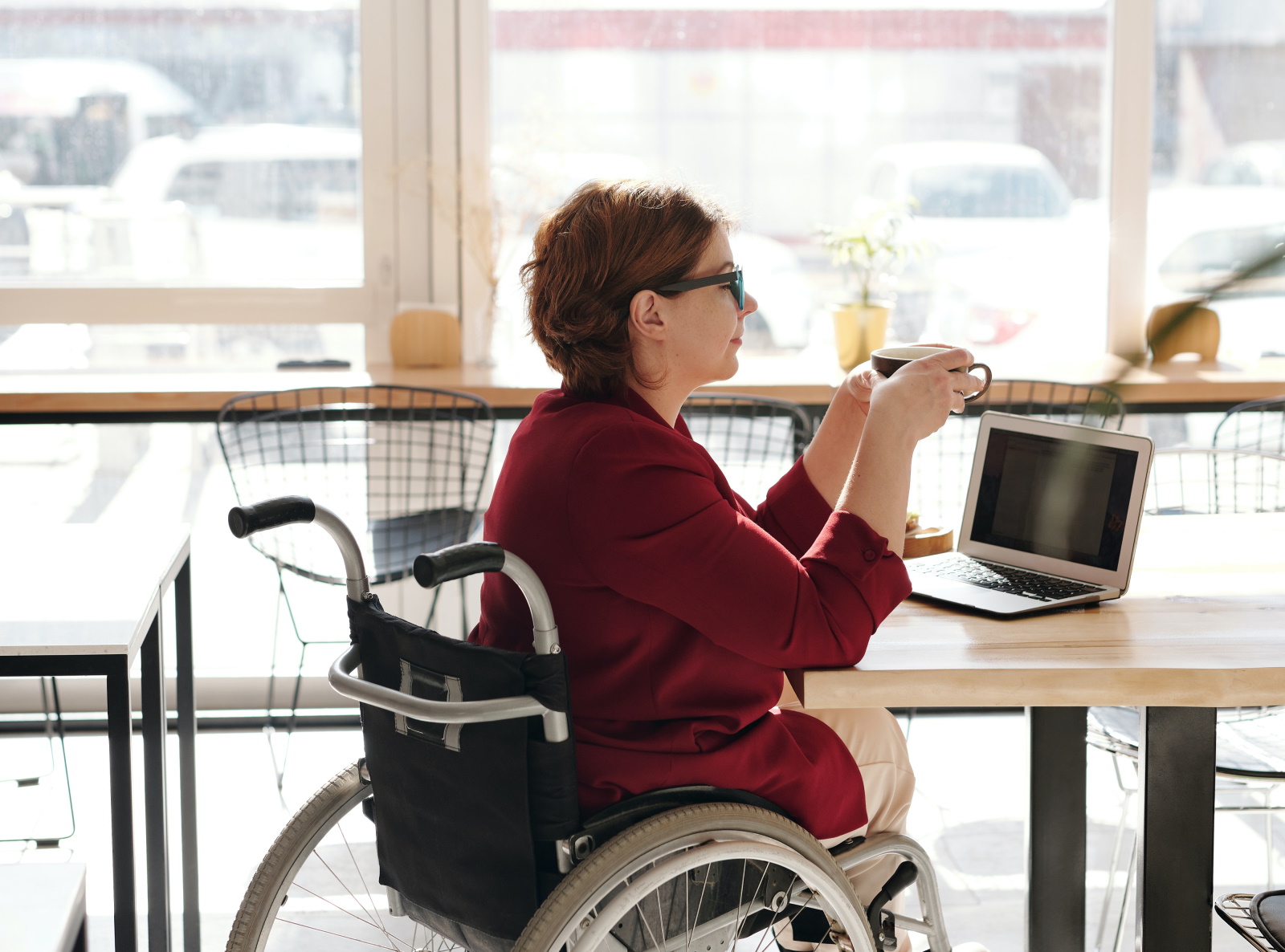 A woman in a wheelchair sat enjoying a cup of coffee as she works in front of a large window